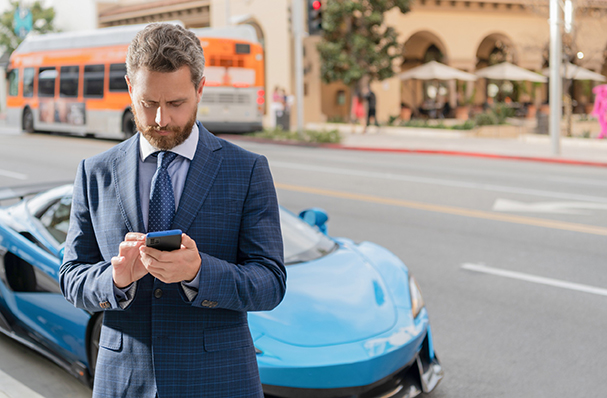Agent immobilier brun en reconversion en costume bleu devant une voiture sportive, focalisé sur son smartphone